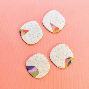 May include: Four speckled, off-white, square-shaped polymer clay earrings with rounded corners. Each earring features a colorful, abstract design in shades of pink, yellow, blue, and green. The earrings are arranged on a light pink background.