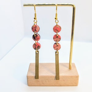 May include: A pair of dangle earrings with gold-colored hooks. Each earring features three round, marbled beads in shades of red, pink, and brown, connected by small gold-colored rings. A rectangular gold-colored bar hangs below the beads.
