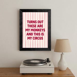 May include: A framed print with the text "Turns out these are my monkeys and this is my circus" in red letters. The background has pink and white vertical stripes. A white record player and lamp are in the foreground.