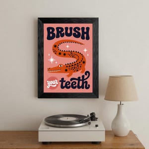 May include: A framed art print with a pink background features an orange alligator illustration. The words "BRUSH your teeth" are in navy blue and white. The print is in a black frame and is displayed on a wooden surface.