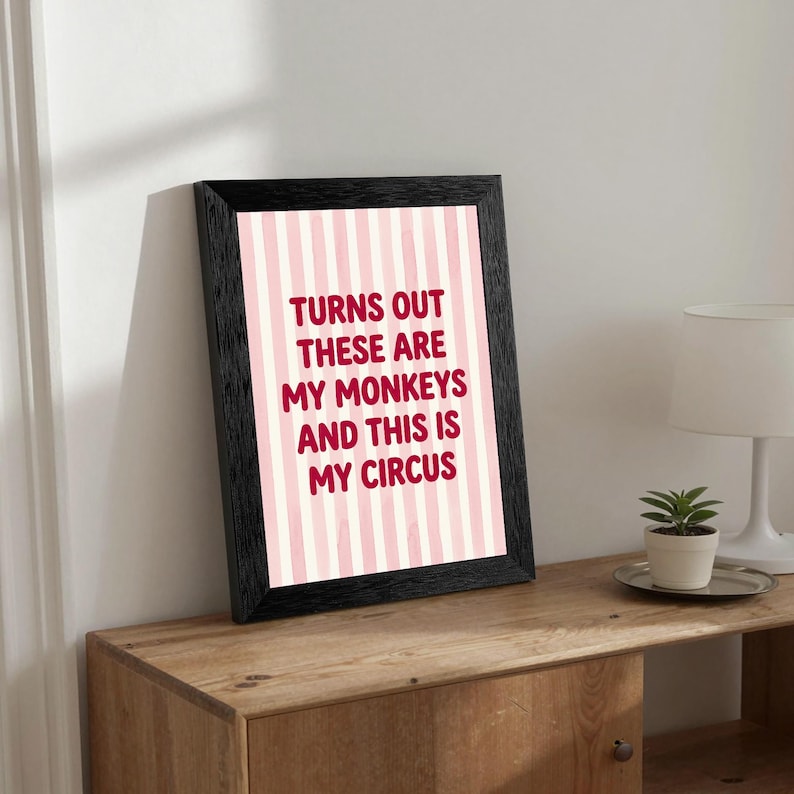 May include: A framed print with the text "Turns out these are my monkeys and this is my circus" in a bold, red font. The background features pink and white vertical stripes. The frame is black and the print is displayed on a wooden surface.