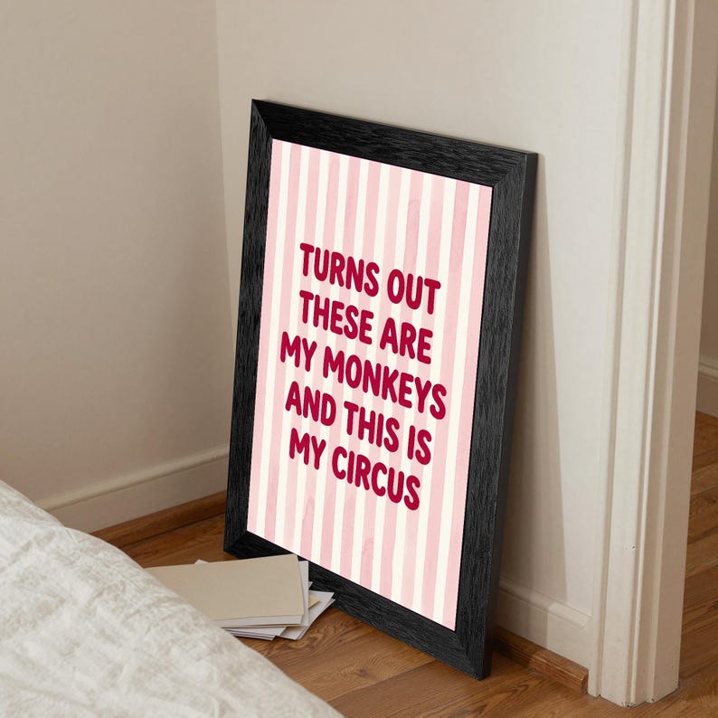 May include: A framed print with a black border, leaning against a wall. The print features a pink and white striped background with the text "TURNS OUT THESE ARE MY MONKEYS AND THIS IS MY CIRCUS" in a bold, red font.