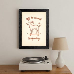 May include: A framed art print with a black frame. The print features a brown line drawing of a dog on a skateboard, with the words "Off to commit Tomfoolery" in brown script. The print is displayed on a wall above a record player and lamp.
