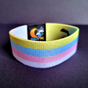 May include: A fabric wristband featuring horizontal stripes in yellow, light blue, pink, and white. A black rectangular tag is attached to the band, displaying a graphic of a crescent moon and the text SOVTAL in white and orange lettering.
