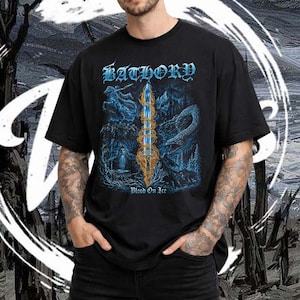 Op de afbeelding: Zwart t-shirt met de bandnaam "Bathory" in blauwe gotische letters en een blauw-gouden illustratie van een zwaard en een draak. De tekst "Blood On Ice" staat eronder. Het t-shirt wordt door een persoon gedragen.
