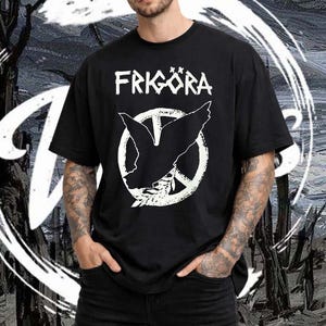 Puede incluir: Camiseta negra con la palabra "FRIGÖRA" en blanco, sobre un símbolo de la paz blanco con una paloma y una rama de olivo. La persona lleva tatuajes en los brazos y va vestida con vaqueros negros.
