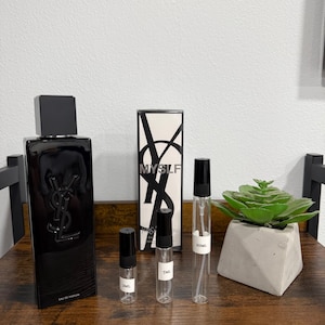 May include: A black perfume bottle with a silver logo and a black cap, next to a box with the word "MYSLF" in white. There are three clear glass vials with black caps and labels indicating 2ml, 5ml, and 10ml. A small succulent plant in a geometric concrete pot is also present.