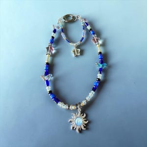May include: A beaded necklace and bracelet set featuring blue, clear, and black beads. The necklace has a silver sun pendant and butterfly charms. The bracelet has a silver star charm. Pink star and blue butterfly charms are also present.