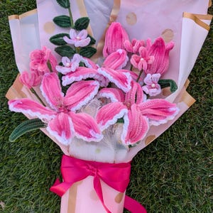 May include: A bouquet of pink and white handmade flowers, wrapped in decorative paper with gold polka dots and a pink ribbon. The flowers include lilies, tulips, and other floral arrangements, with green stems and leaves.