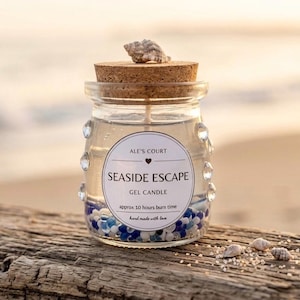 Puede incluir: Vela de gel "Seaside Escape" en un tarro de cristal con tapa de corcho y una concha. El tarro está decorado con gemas transparentes y contiene cuentas azules y blancas. Duración de la combustión de aproximadamente 10 horas.