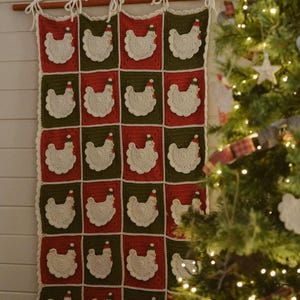 May include: A rectangular crochet wall hanging featuring a grid of alternating red and dark green squares. Each square displays a white crocheted chicken applique wearing a small red and green hat. The piece is suspended from a wooden rod.