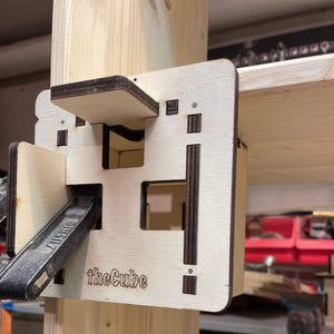 Puede incluir: Un sistema de soporte de madera, con un diseño cuadrado con secciones recortadas y el texto "theCube". Una abrazadera metálica está unida al soporte, asegurándolo a un poste y estante de madera. El soporte es de madera clara.