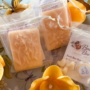 May include: Close-up of handmade soaps in clear bags, with floral designs. The soaps are a light orange color. The image also shows a small jar of "Bee Balm" and a package with the text "Bare Bee Botanicals". The background includes yellow flower petals.
