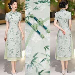 Può includere: Un abito midi verde chiaro con colletto alla coreana, maniche corte e chiusura laterale con due bottoni sferici. Il tessuto presenta un motivo jacquard floreale bianco sovrapposto a una stampa di foglie di bambù verdi.