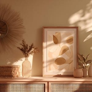May include: A serene interior scene featuring a wooden cabinet with decorative items. A framed abstract art piece, vases with dried flowers, and a potted plant with large green leaves are arranged on the cabinet. The room is bathed in warm sunlight.