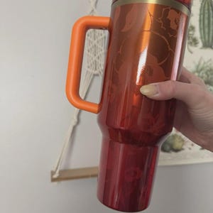May include: A red and orange travel mug with a floral pattern. The mug has a large orange handle and a matching orange lid. The body of the mug is a deep red with a metallic sheen, and the floral design is a lighter shade of red.
