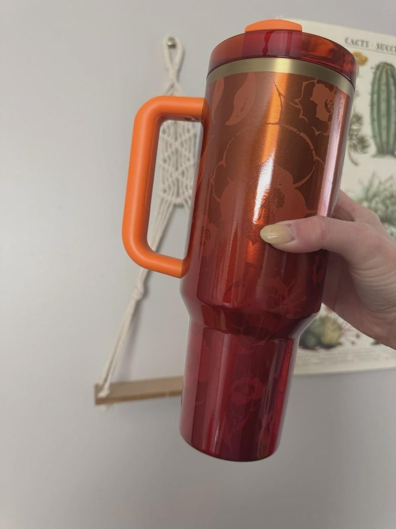 May include: A red and orange travel mug with a floral pattern. The mug has a large orange handle and a matching orange lid. The body of the mug is a deep red with a metallic sheen, and the floral design is a lighter shade of red.