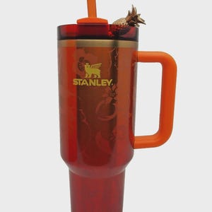 May include: A red and orange Stanley tumbler with a handle and straw. The tumbler has a gold logo and a decorative pineapple on the lid. The tumbler is designed with a floral pattern.