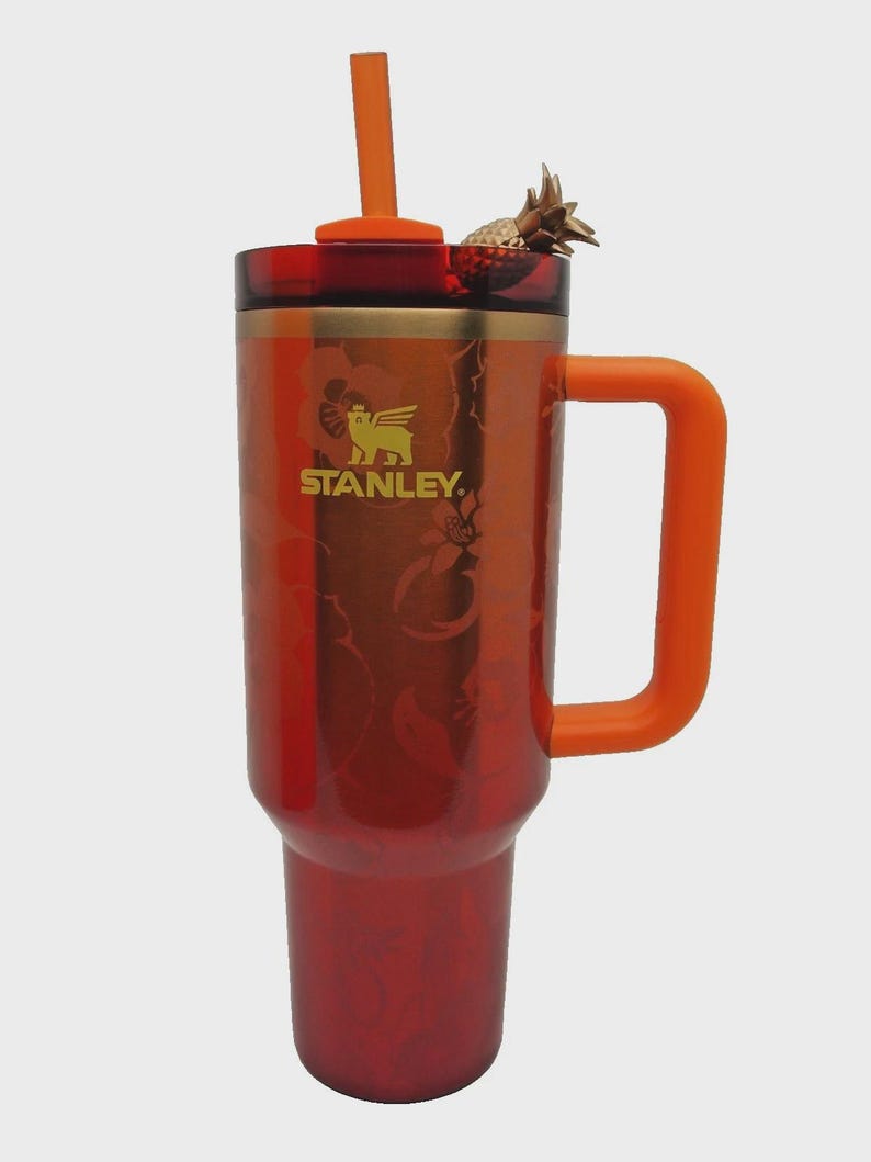 May include: A red and orange Stanley tumbler with a handle and straw. The tumbler has a gold logo and a decorative pineapple on the lid. The tumbler is designed with a floral pattern.