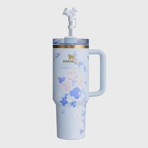 May include: A pale blue Stanley tumbler with a floral pattern of pink and blue flowers. It has a handle, a clear lid, and a white bow-shaped straw topper. The text "STANLEY" and "LOVESHACKFANCY" are printed on the tumbler. A stylish drinkware item.
