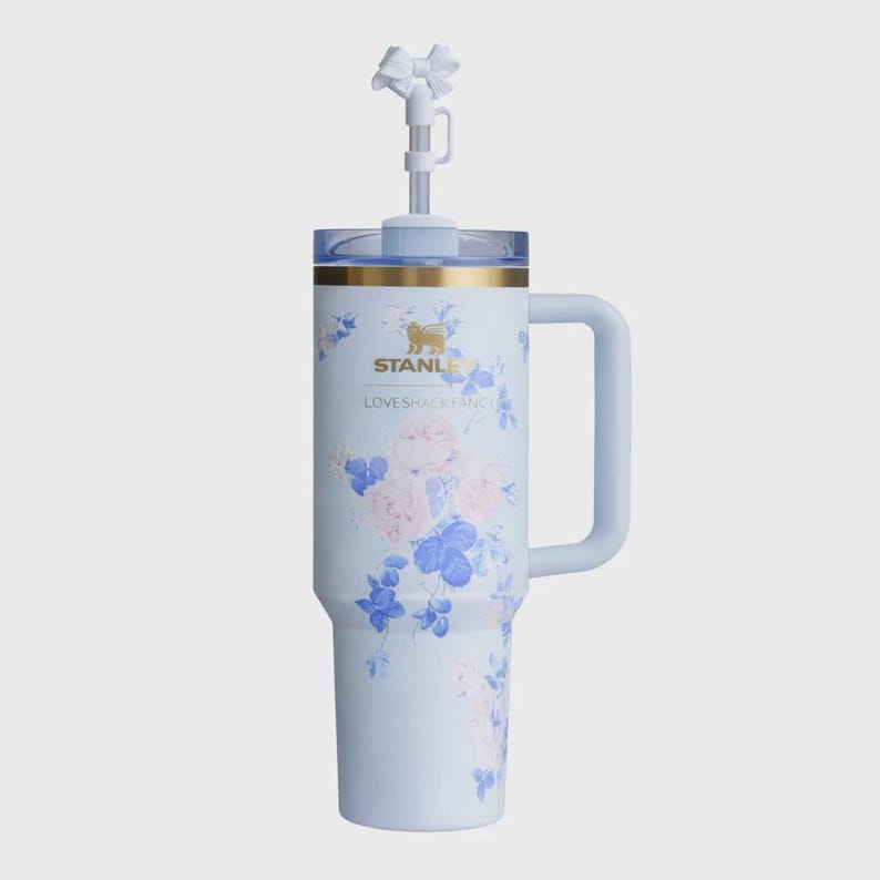 May include: A pale blue Stanley tumbler with a floral pattern of pink and blue flowers. It has a handle, a clear lid, and a white bow-shaped straw topper. The text "STANLEY" and "LOVESHACKFANCY" are printed on the tumbler. A stylish drinkware item.
