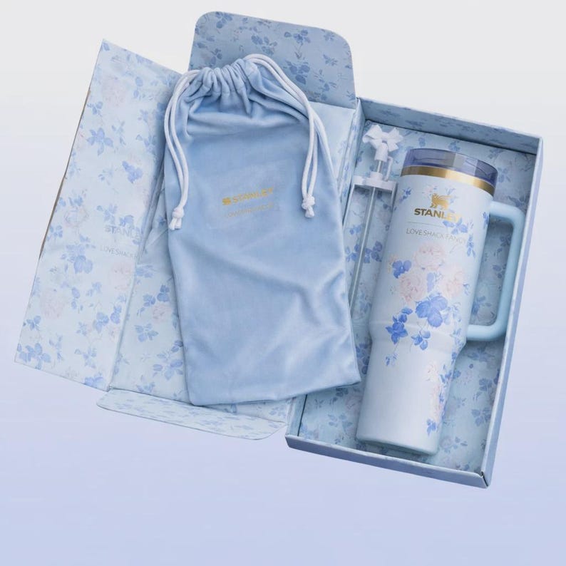 May include: A light blue Stanley tumbler with a handle, floral design, and a straw, presented in a floral-patterned box with a matching drawstring bag. The tumbler has the text "STANLEY LOVE SHACK FANS".