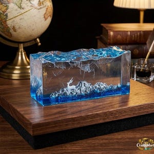 May include: A rectangular resin art piece featuring a miniature landscape. The clear resin block has a blue wave design on top, with a white deer and snow-capped mountains inside. The piece sits on a wooden surface.