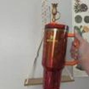 May include: A red and orange insulated travel mug with a handle. The mug has a metallic finish and a matching lid. The handle is orange. The mug is being held in front of a white wall.