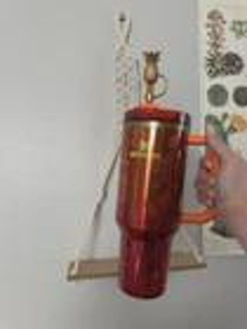 May include: A red and orange insulated travel mug with a handle. The mug has a metallic finish and a matching lid. The handle is orange. The mug is being held in front of a white wall.