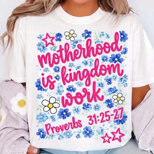 Puede incluir: Camiseta blanca con el texto rosa "Motherhood is kingdom work", rodeada de diseños florales azules y blancos y estrellas. El versículo bíblico Proverbios 31:25-27 también está impreso en la camiseta.