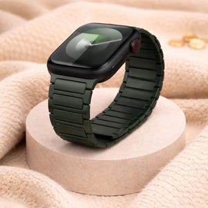 May include: A black smartwatch with a dark green metal band, displayed on a small, round, beige pedestal. The watch face is black with a green and white display. The band is made of rectangular links.