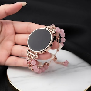 May include: Rose gold smartwatch with a black screen and a pink beaded bracelet. The watch has a round face and is held in a hand. The bracelet features small, round pink beads and a delicate chain.