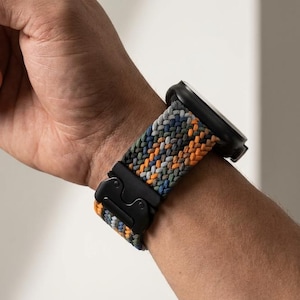 May include: A black smartwatch with a multi-colored braided band in shades of orange, green, blue, and gray. The band is secured with a black clasp. The watch is on a wrist.