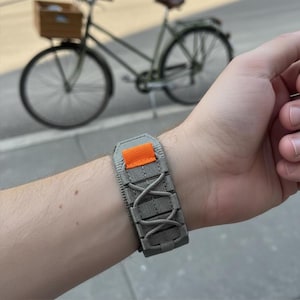 Pode incluir: Uma pulseira tática cinza e laranja está presa no pulso de uma pessoa. A pulseira apresenta uma base cinza com um detalhe laranja e um design cruzado. Uma bicicleta é visível ao fundo.