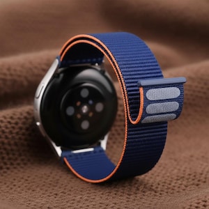 Puede incluir: Un reloj inteligente plateado con una correa de nailon azul marino y naranja. La correa tiene un tejido texturizado y un cierre gris. La esfera del reloj es negra con detalles plateados.