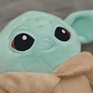 May include: A plush toy of a character with large black eyes, a light green face, and large ears. The toy has a tan robe and a small embroidered smile. The background is a solid gray.
