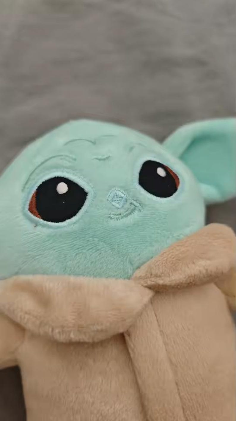 May include: A plush toy of a character with large black eyes, a light green face, and large ears. The toy has a tan robe and a small embroidered smile. The background is a solid gray.