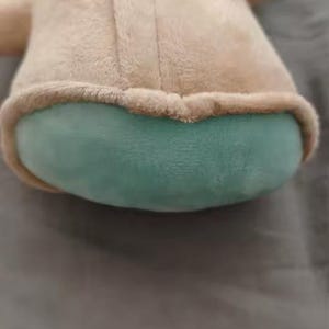 May include: A plush toy depicting a character with large ears, a tan coat, and a teal-colored face and accents. The toy has a rounded body and is made of soft, fuzzy material.