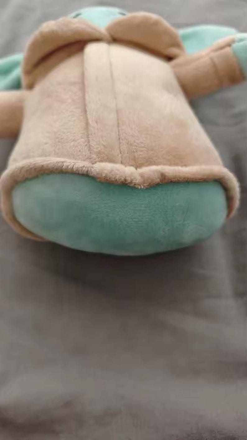 May include: A plush toy depicting a character with large ears, a tan coat, and a teal-colored face and accents. The toy has a rounded body and is made of soft, fuzzy material.