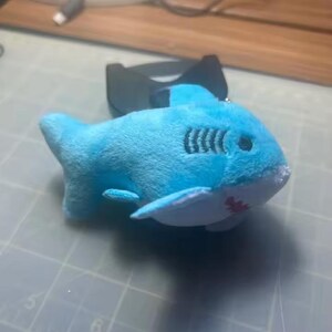 May include: A plush toy shark in shades of blue, with white accents on the fins and belly. The shark has black stitched gills and a red mouth. The toy is lying on a gray surface.