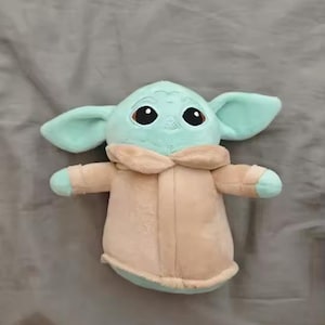 May include: A plush toy of a character with large ears, big eyes, and a tan coat. The toy is light green and tan, and is lying on a gray surface. The toy is a popular character from a science fiction franchise.