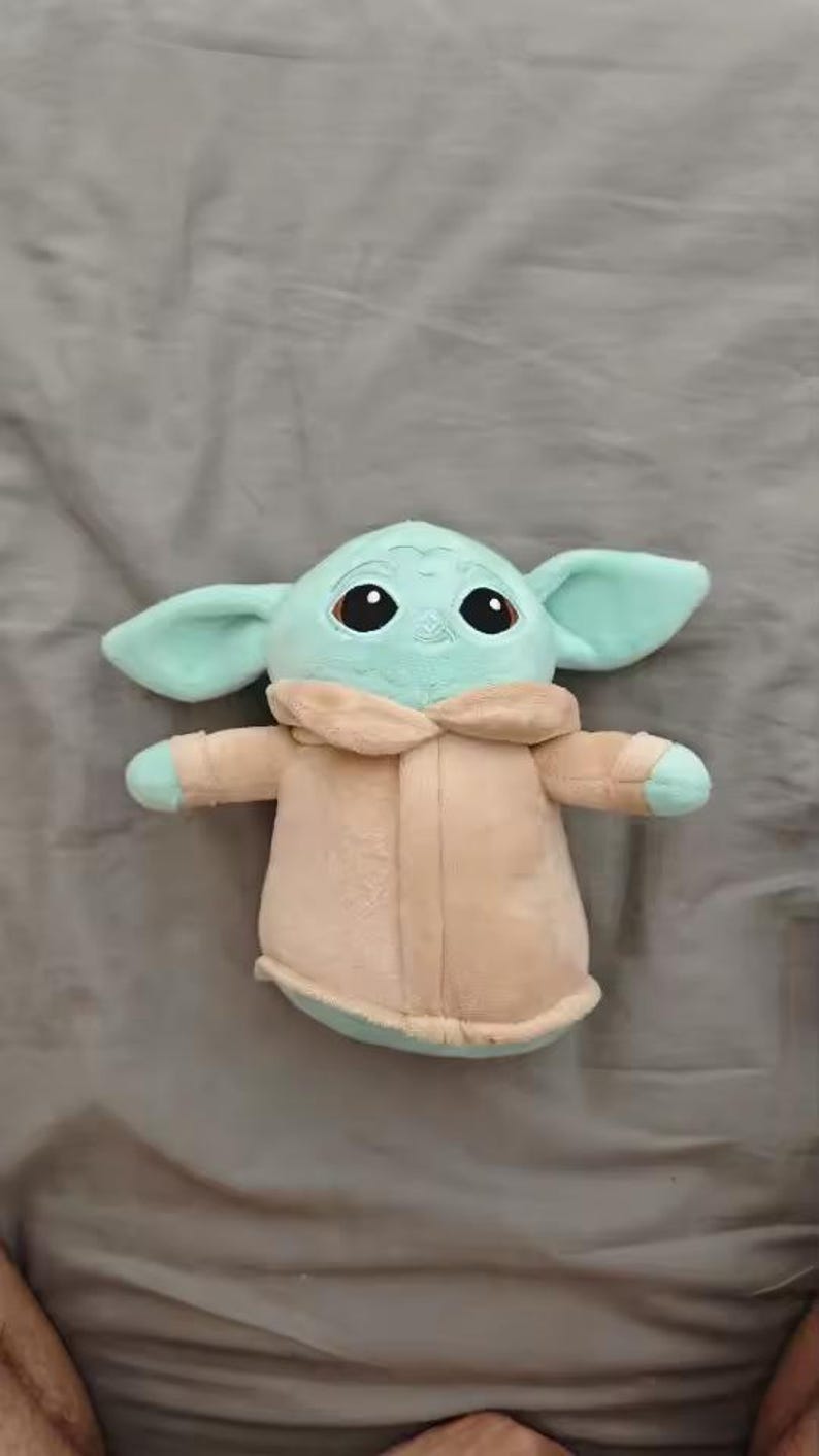 May include: A plush toy of a character with large ears, big eyes, and a tan coat. The toy is light green and tan, and is lying on a gray surface. The toy is a popular character from a science fiction franchise.