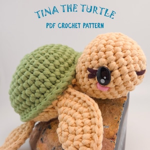 Op de afbeelding: Een gehaakte schildpad met een groene schild en een bruin lichaam. De schildpad heeft grote zwarte ogen, een roze mond en zit op een steen. De tekst "TINA THE TURTLE PDF CROCHET PATTERN" staat boven de schildpad.