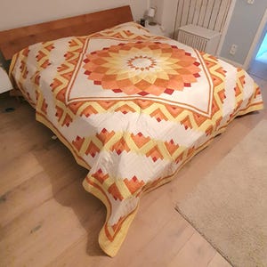 Op de afbeelding: Een handgemaakte quilt met een geometrisch ontwerp in tinten oranje, geel en rood op een witte achtergrond. De quilt is over een houten bedframe verspreid. Het patroon van de quilt heeft een centraal cirkelvormig ontwerp en herhaalde geometrische vormen.