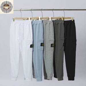 Può includere: Sei paia di pantaloni della tuta in vari colori, tra cui bianco, azzurro, grigio, grigio scuro e nero, sono appesi a un attaccapanni. Ogni paio presenta una tasca cargo e una piccola toppa circolare con logo sulla gamba sinistra. I pantaloni hanno una vita con coulisse e polsini alle caviglie.