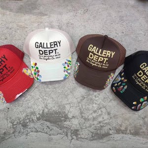 May include: Four trucker hats with the text "GALLERY DEPT." in bold letters. The hats are red, white, brown, and black. Each hat has a mesh back and a curved brim. The text "7513 Beverly Blvd Los Angeles Ca." is printed below the main text.