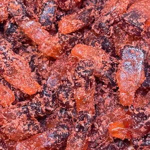 May include: Close-up of a pile of red and dark purple gummy candies. The candies have a cola bottle shape and are coated in a fine, red sugar. The image shows a close-up view of the candies.