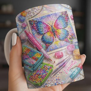 May include: White ceramic mug featuring a colorful butterfly design, surrounded by craft supplies. The design includes a pink pen, containers of beads, and the name "Emma". The mug is ideal for crafters.