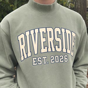 May include: A sage green mock neck sweatshirt featuring the text RIVERSIDE in a large, arched, cream-colored font with a dark blue outline, positioned above the text EST. 2026 in a smaller, matching cream and dark blue font on the chest.