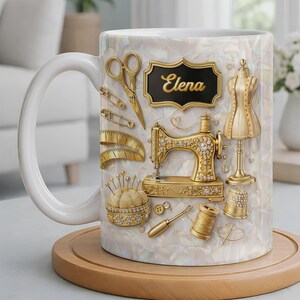 May include: White ceramic mug featuring a gold sewing machine, scissors, pins, and a tailor's dummy. The mug has a pearlescent background and the name "Elena" in gold lettering on a black plaque. A wooden coaster supports the mug.